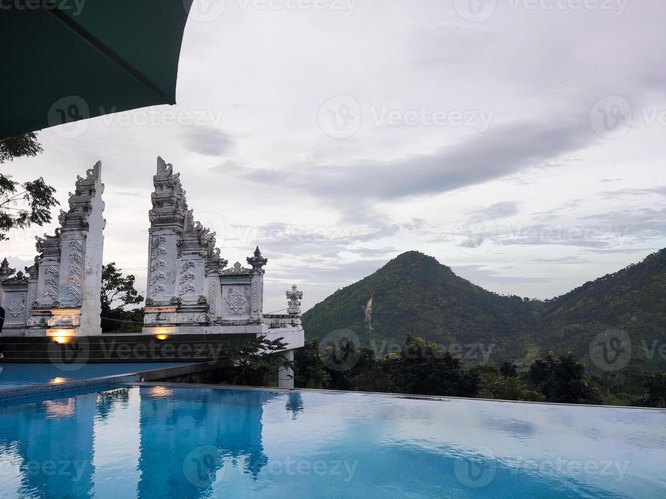 a swimming pool with a clear blue water. In the background, there is a traditional Balinese temple with multiple tiers. There are palm trees and other lush vegetation surrounding the pool and temple. photo