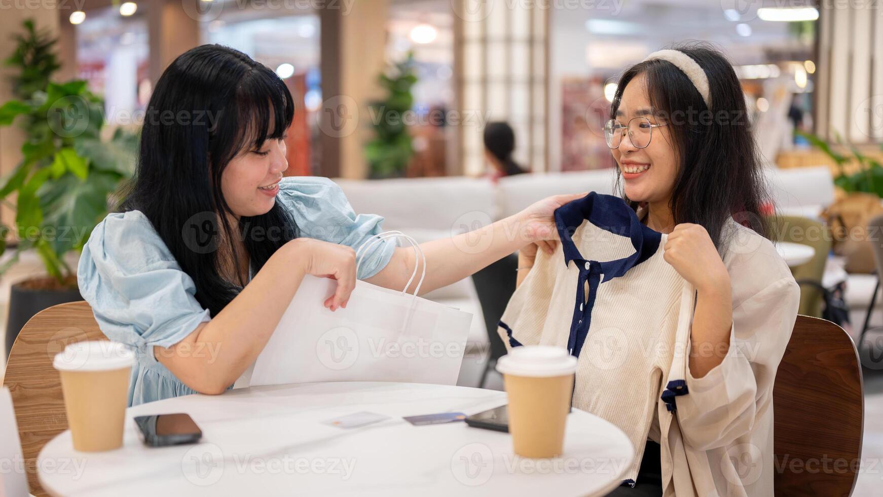 Two Asian women sit in a cafe at a mall, one excitedly showing off a new shirt to her friend. photo
