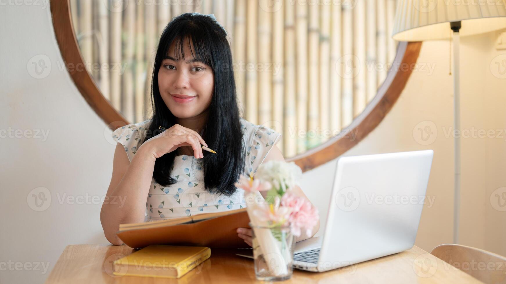 A cute Asian woman sits at a table in a coffee shop with her laptop computer, smiling at the camera. photo