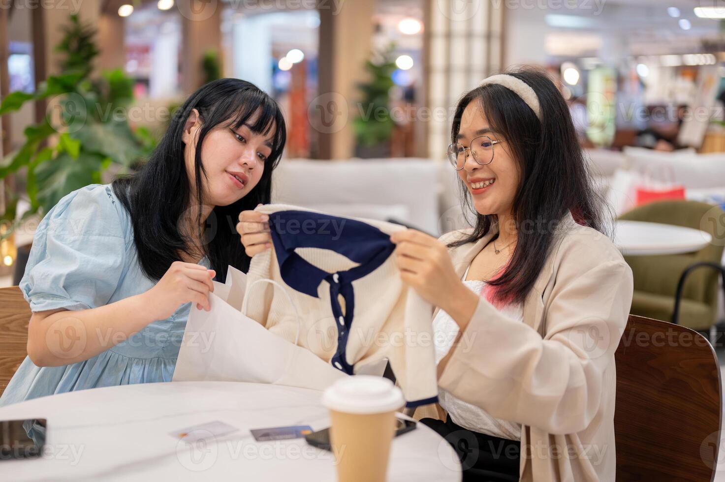 Two young Asian women sit in a cafe at a shopping mall, examining clothes they've purchased. photo
