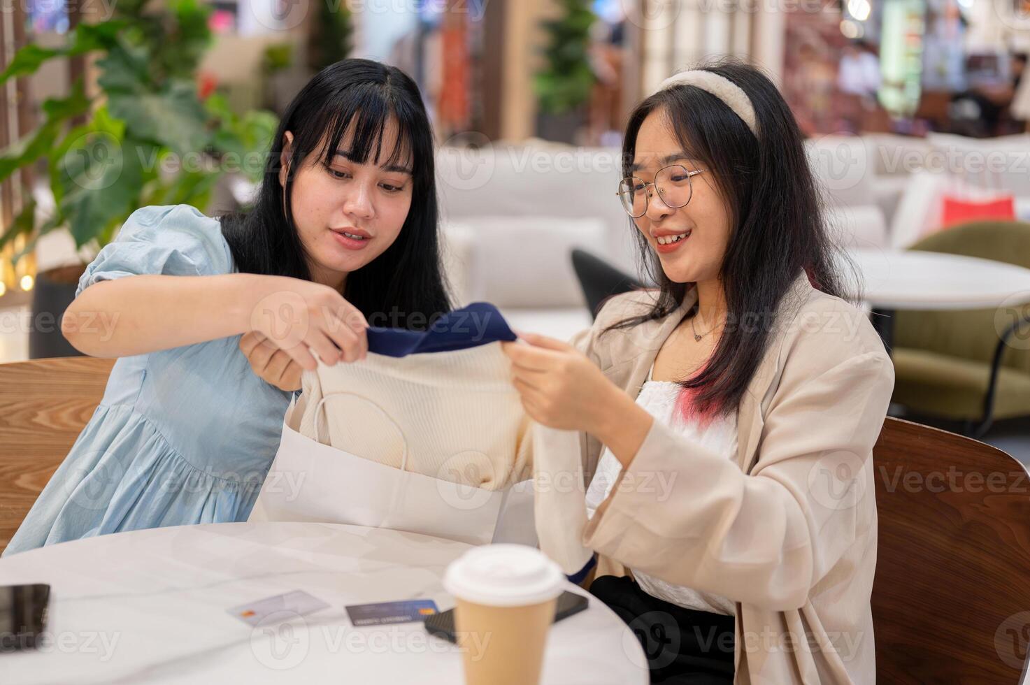 Two young Asian women sit in a cafe at a shopping mall, examining clothes they've purchased. photo