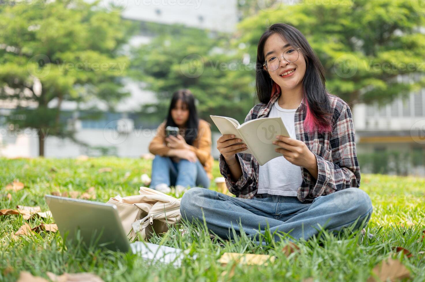 A smiling Asian female student is reading a book while sitting on the grass in a campus park. photo