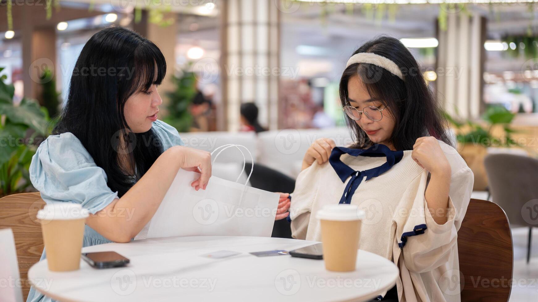 Two Asian women sit in a cafe at a mall, one excitedly showing off a new shirt to her friend. photo