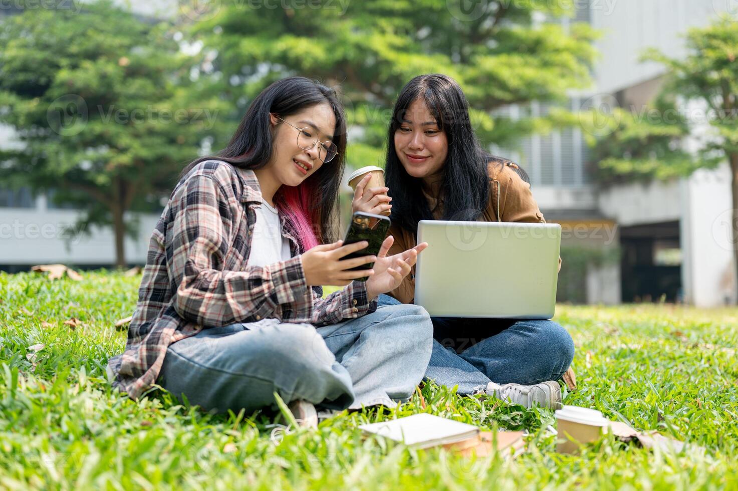 Two college students are sitting on grass in a park, discussing something on a smartphone together. photo