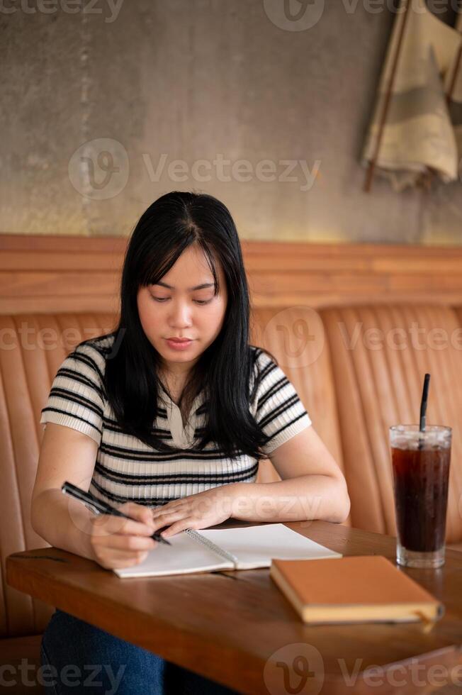 A young Asian woman is writing her diary or taking notes in her notebook at a coffee shop. photo