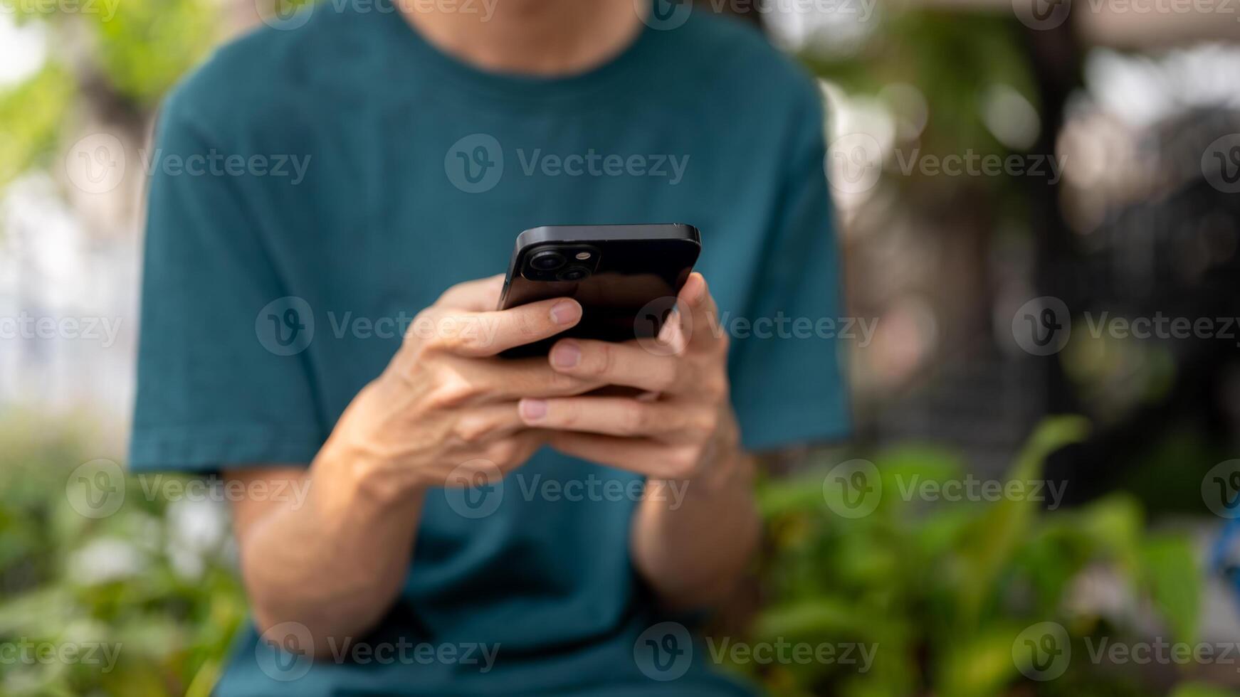 A cropped image of a man in casual wear using his smartphone outdoors, sitting on a bench in a park. photo