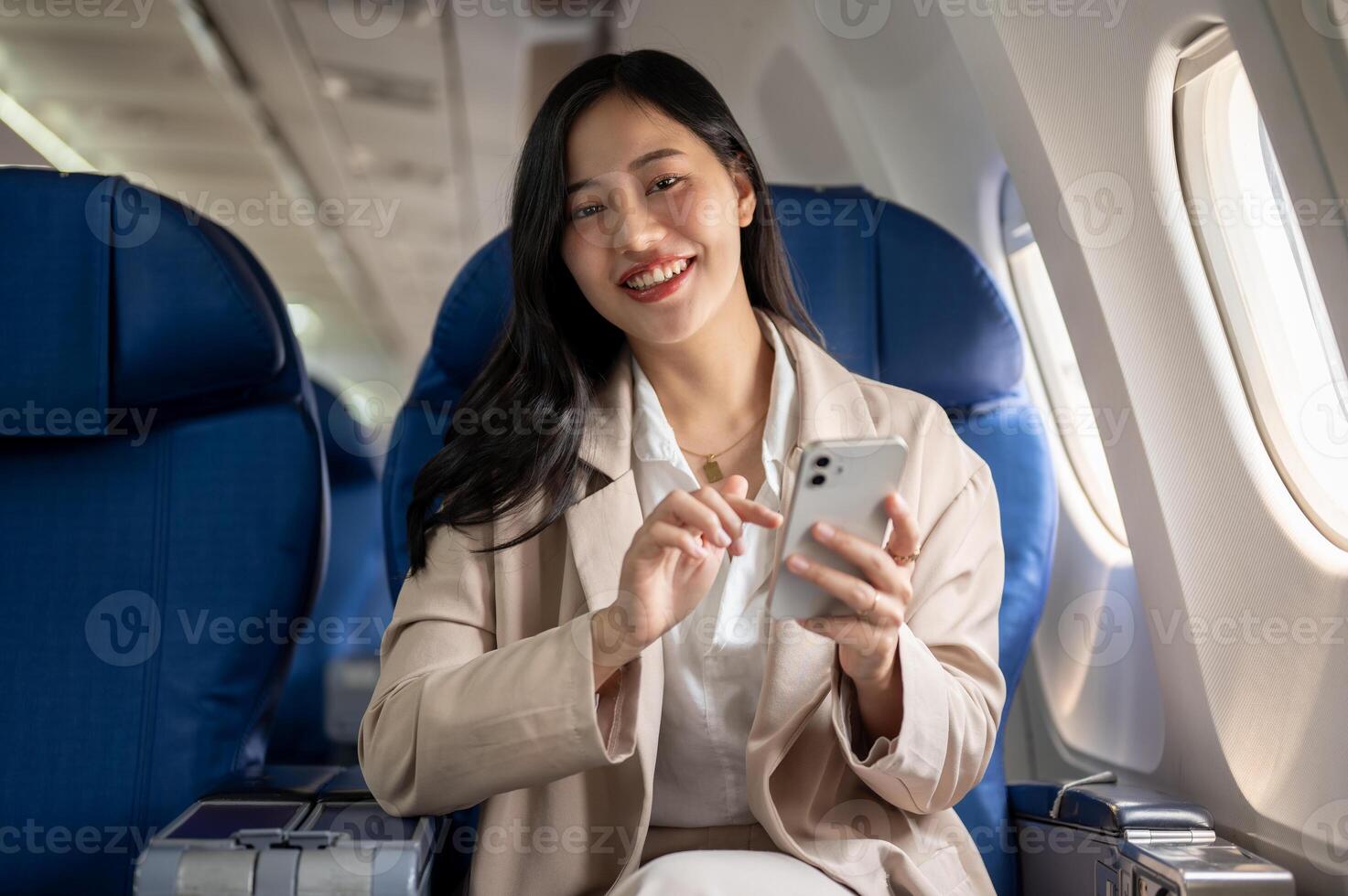 un atractivo asiático mujer de negocios es sentado a el ventana asiento con un teléfono inteligente en su mano. foto