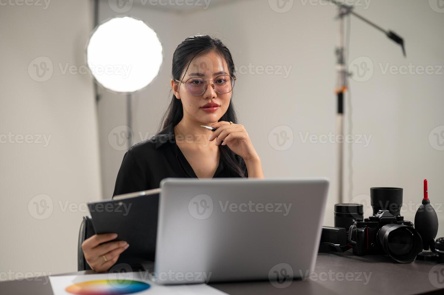 An attractive Asian female photographer is looking at the camera while sitting at a desk in a studio photo