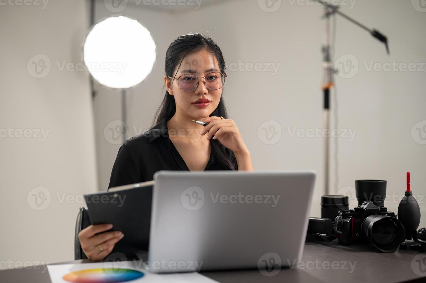 un atractivo asiático hembra fotógrafo es mirando a el cámara mientras sentado a un escritorio en un estudio foto