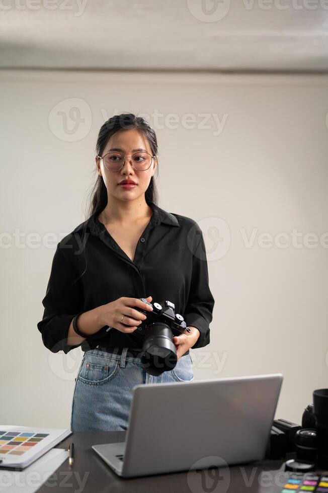 un elegante asiático hembra fotógrafo en un negro camisa con su dslr cámara es en pie en su estudio foto