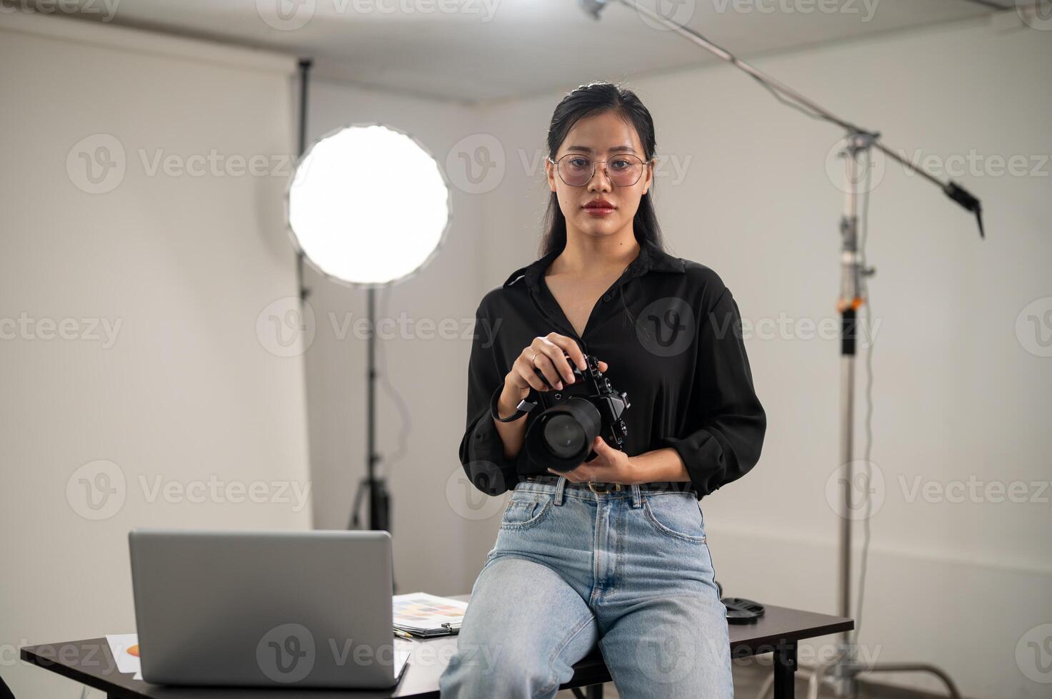 A confident Asian female photographer is holding her DSLR camera, sitting on a desk in a studio. photo