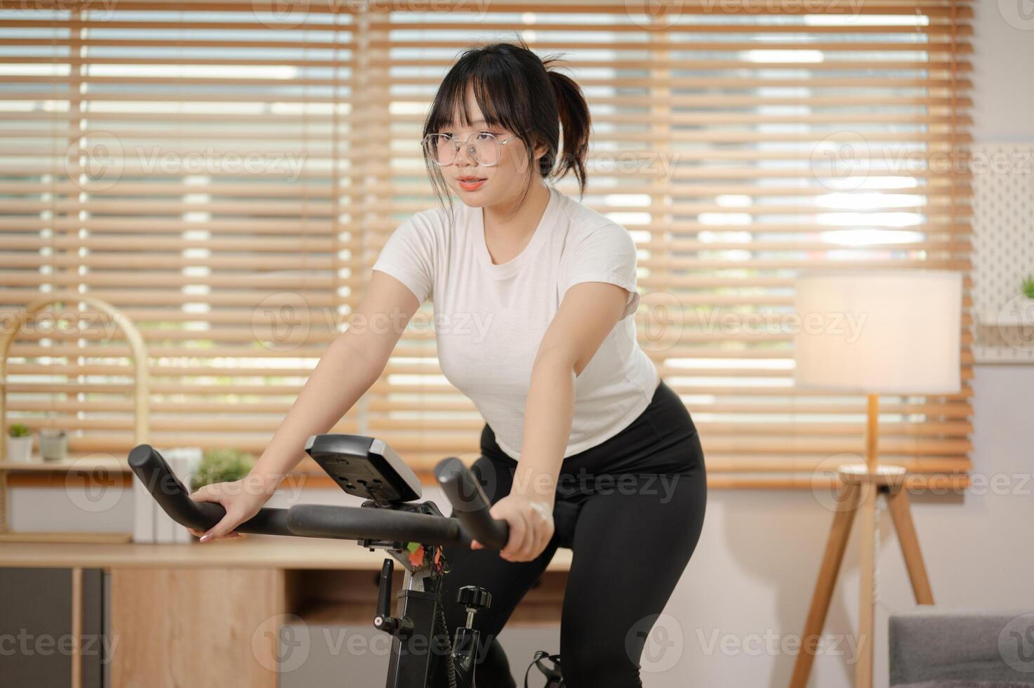 A strong Asian woman in sportswear is exercising on a fitness bike, doing indoor cardio at home. photo