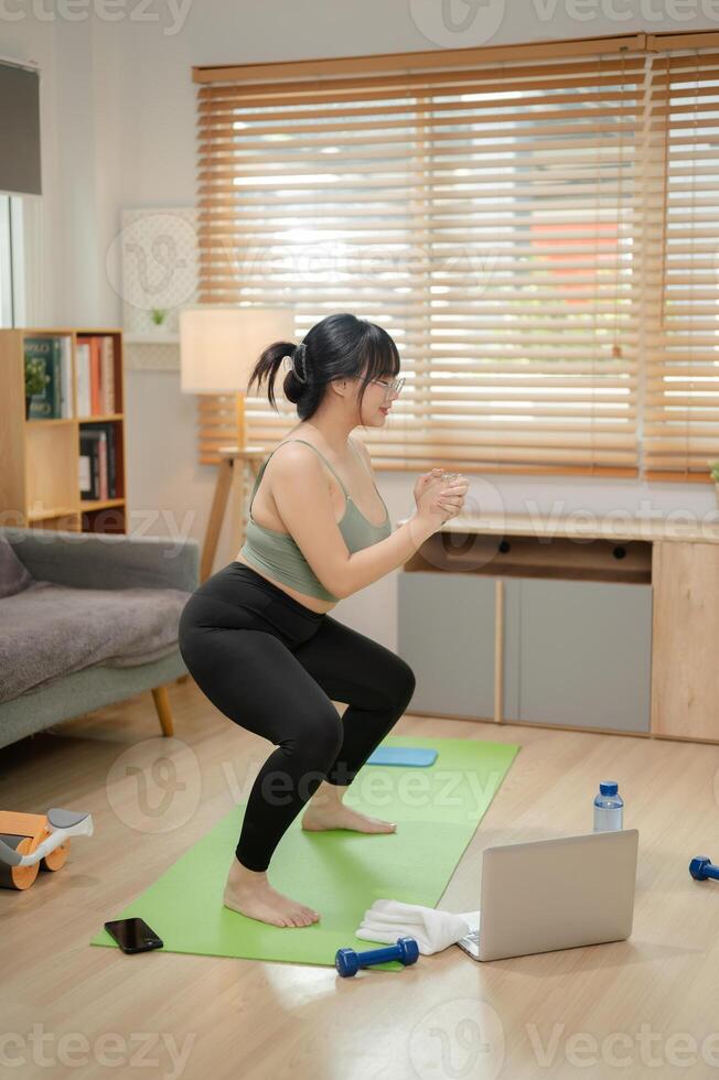 A healthy, strong Asian woman in sportswear is doing a squad on a yoga mat, working out at home. photo