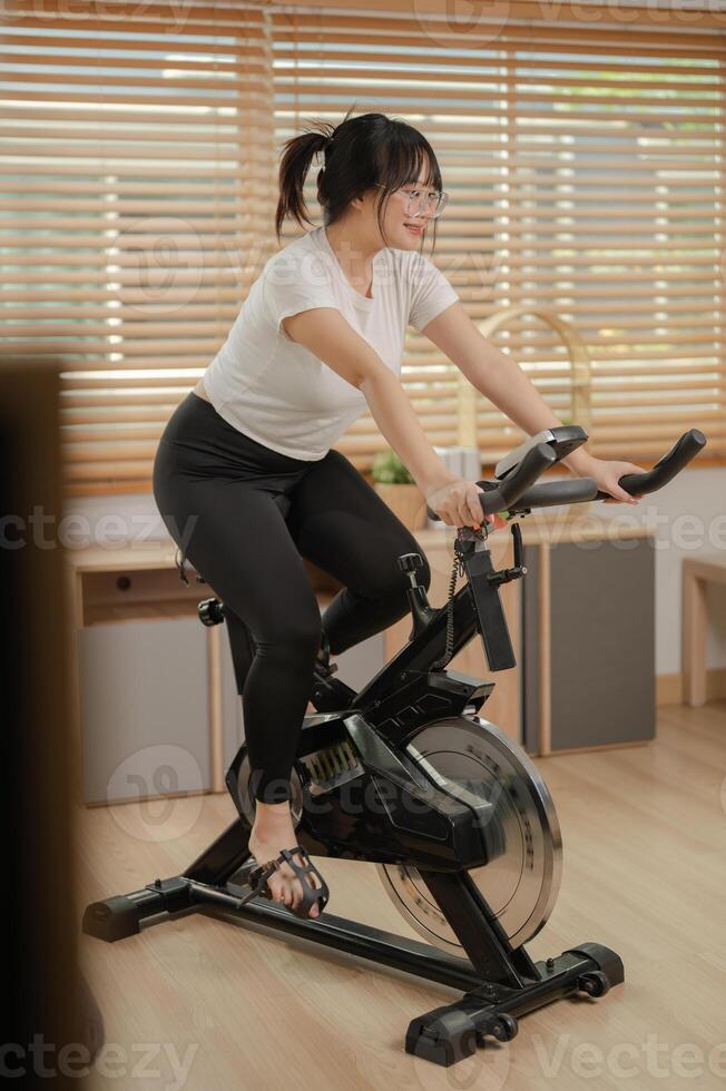 un saludable, ajuste asiático mujer en ropa de deporte es haciendo cardio a hogar, hacer ejercicio en un aptitud bicicleta. foto