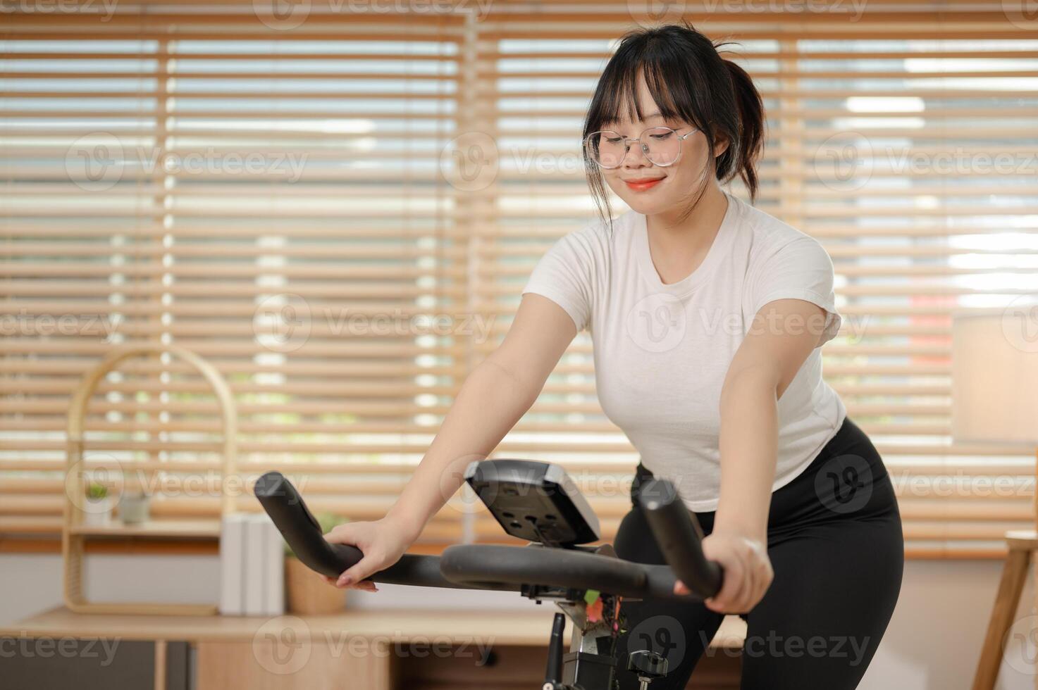 un fuerte asiático mujer en ropa de deporte es hacer ejercicio en un aptitud bicicleta, haciendo interior cardio a hogar. foto