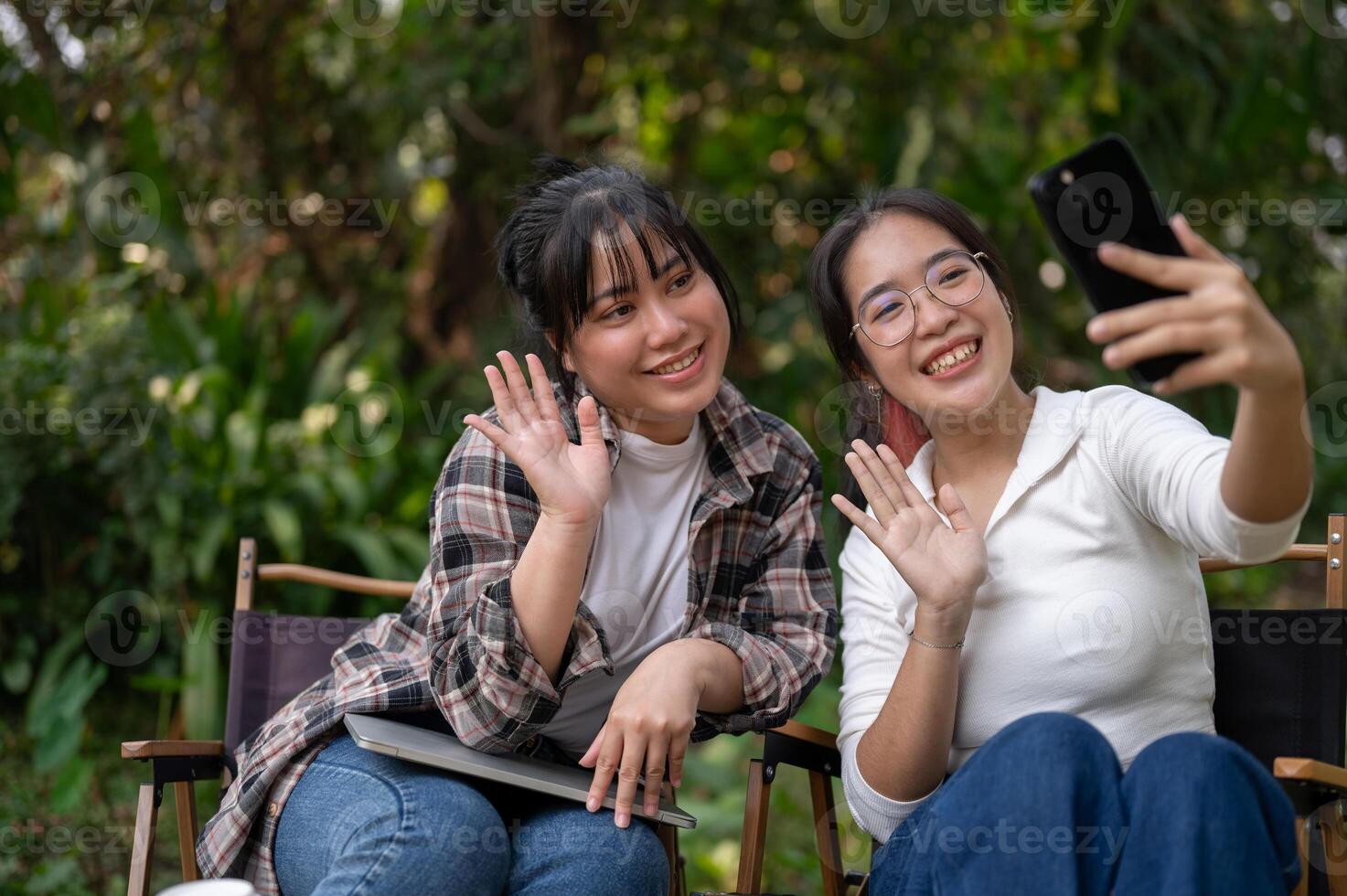 Two cheerful Asian female friends enjoy taking selfies with a smartphone together outdoors. photo