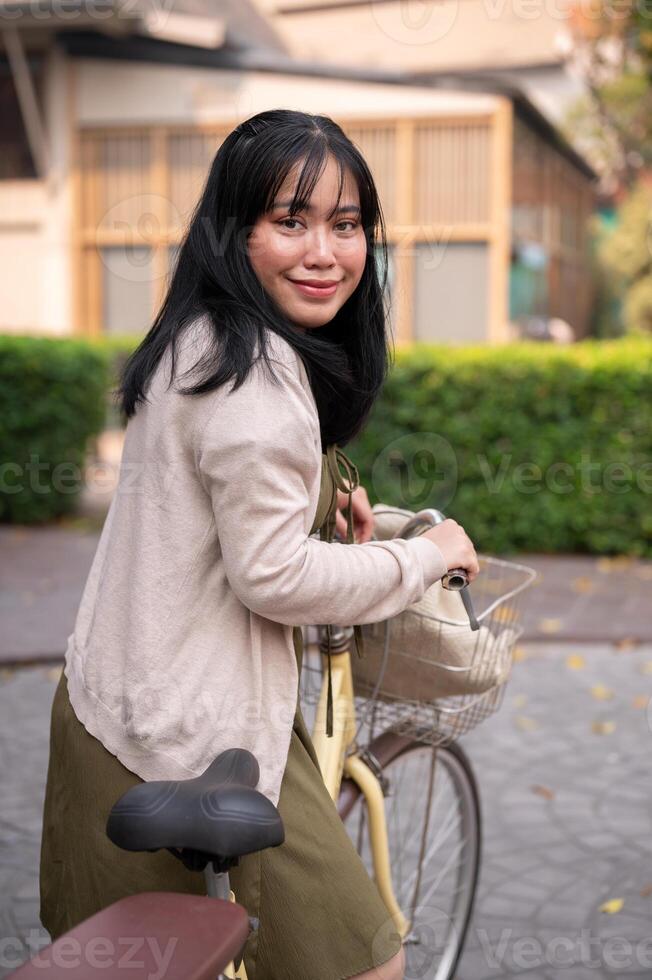 A smiling, happy young Asian woman in a cute dress is riding a bicycle in the city photo