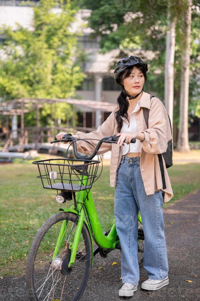 un joven asiático hembra Universidad estudiante vistiendo un bicicleta casco es emprendedor su bicicleta en un verde parque. foto