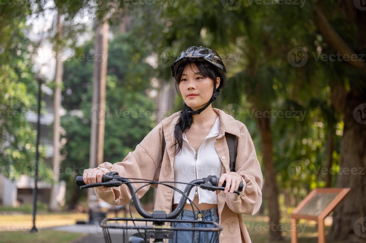 un joven asiático hembra Universidad estudiante vistiendo un bicicleta casco es emprendedor su bicicleta en un verde parque. foto