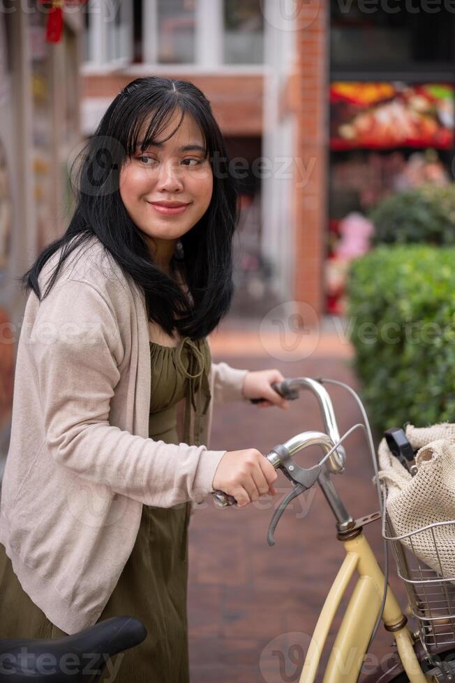 A beautiful, positive Asian woman in a cute dress is walking and pushing her bicycle in the city. photo