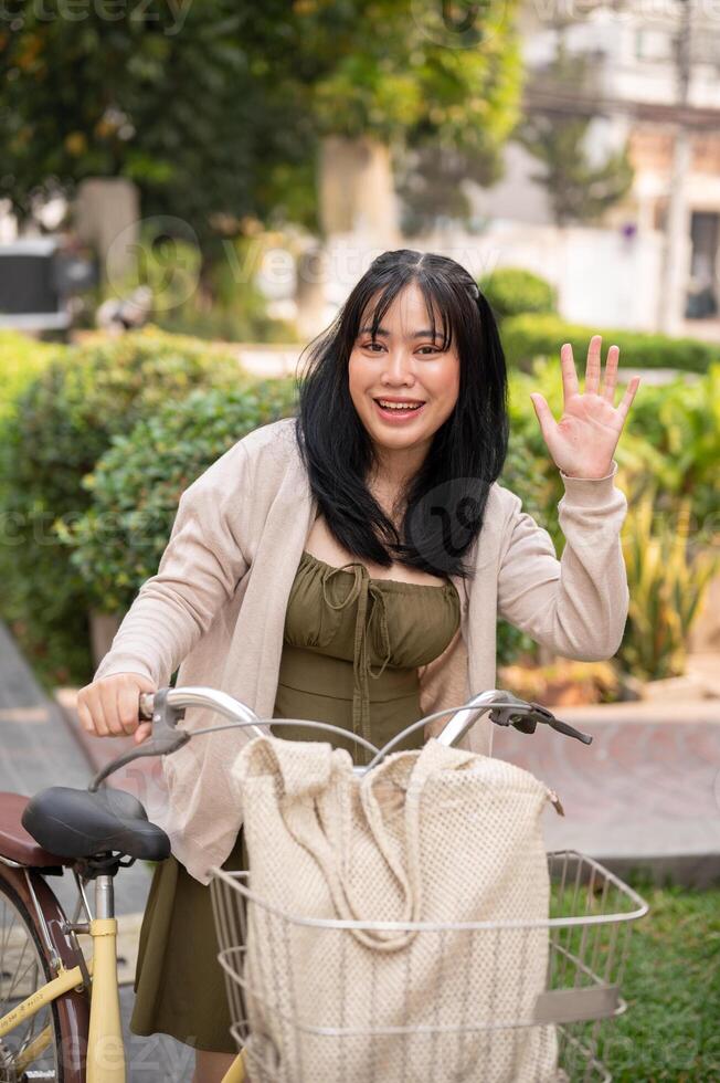 un simpático asiático mujer olas su mano a decir Hola a su amigo mientras emprendedor su bicicleta en el ciudad. foto