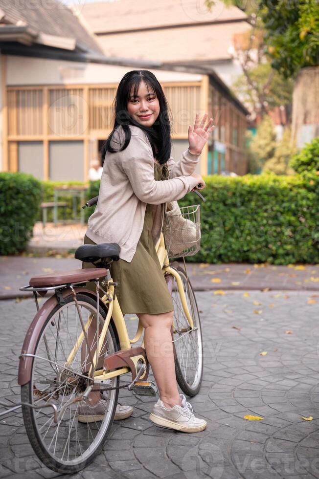 An attractive, smiling Asian woman waves her hand to greet someone while riding a bike in the city. photo