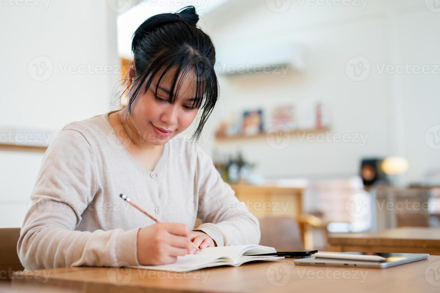 A positive Asian woman is writing in a book, working on her tasks while sitting in a coffee shop. photo