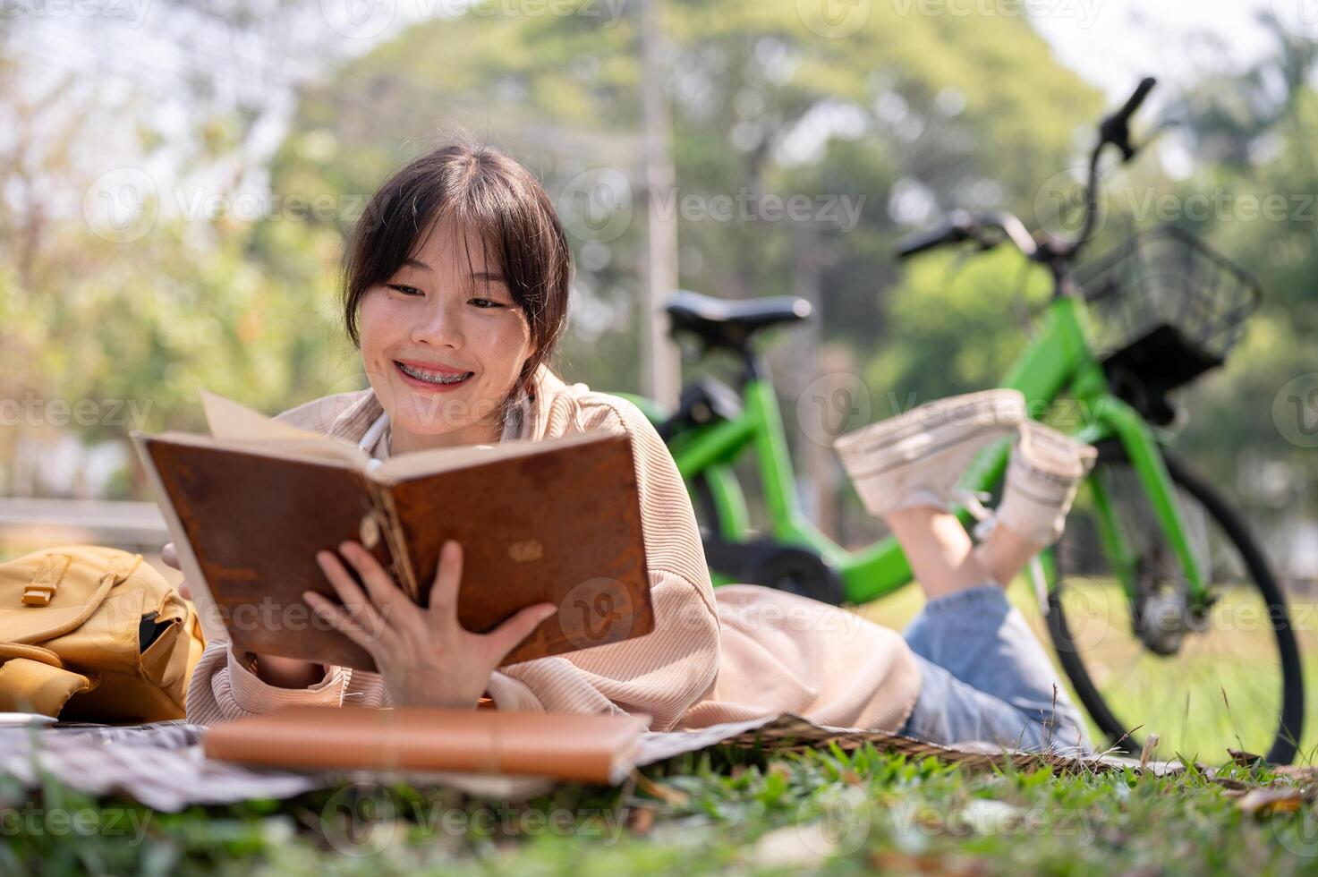 A happy Asian woman is enjoying reading a book on a picnic cloth, while chilling in a green park. photo