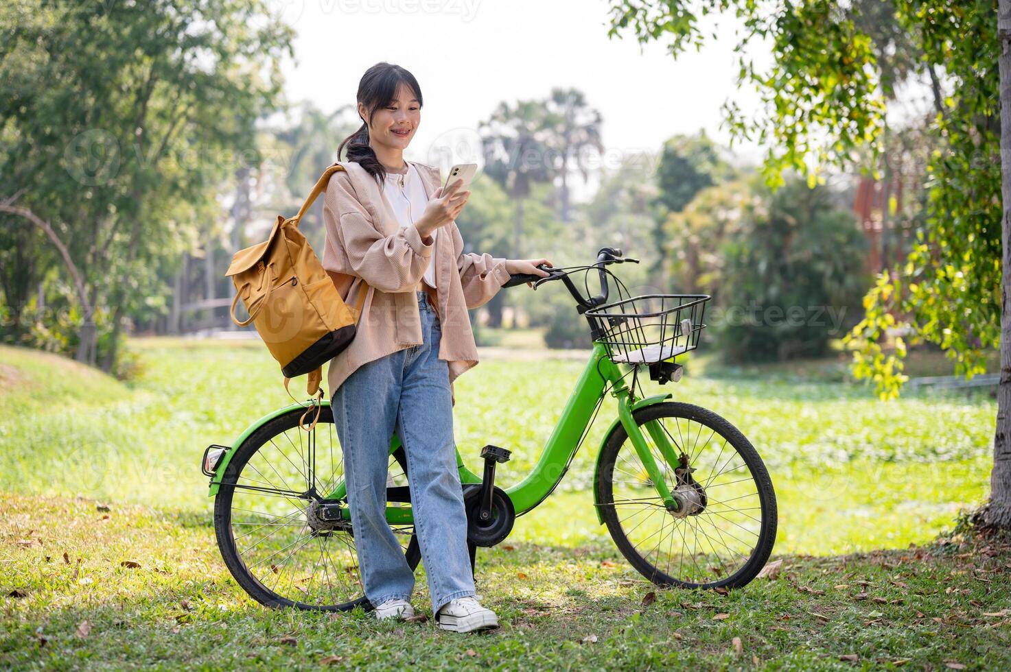 A beautiful Asian woman is using her smartphone while walking with her bike in a sunny green park. photo