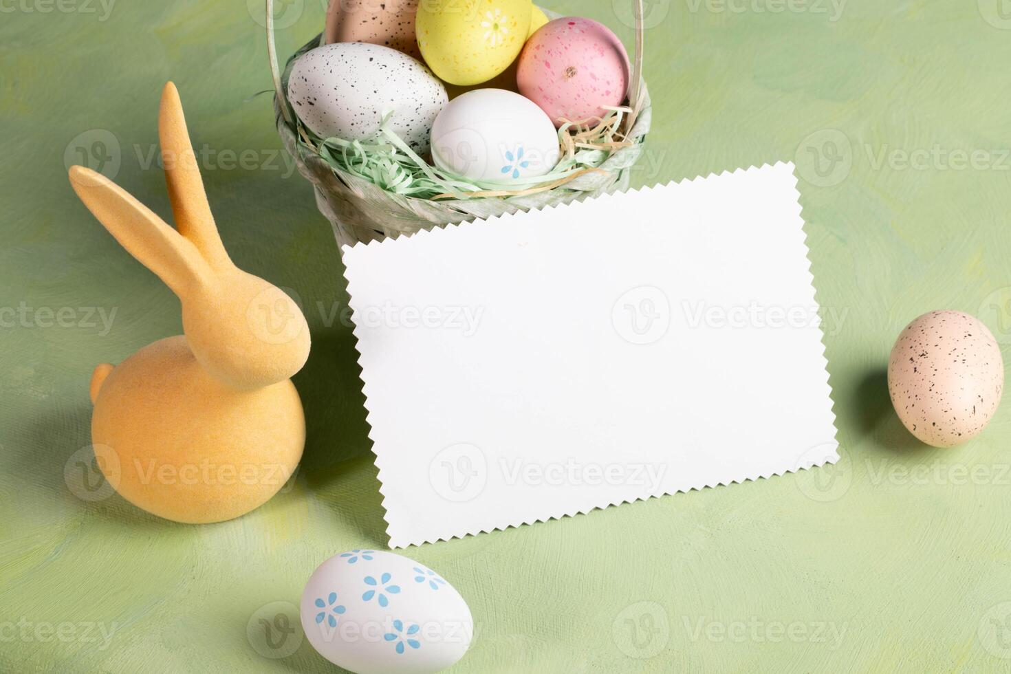 White blank note on pastel yellow green backdrop surrounded by color Easter eggs and yellow bunny. photo