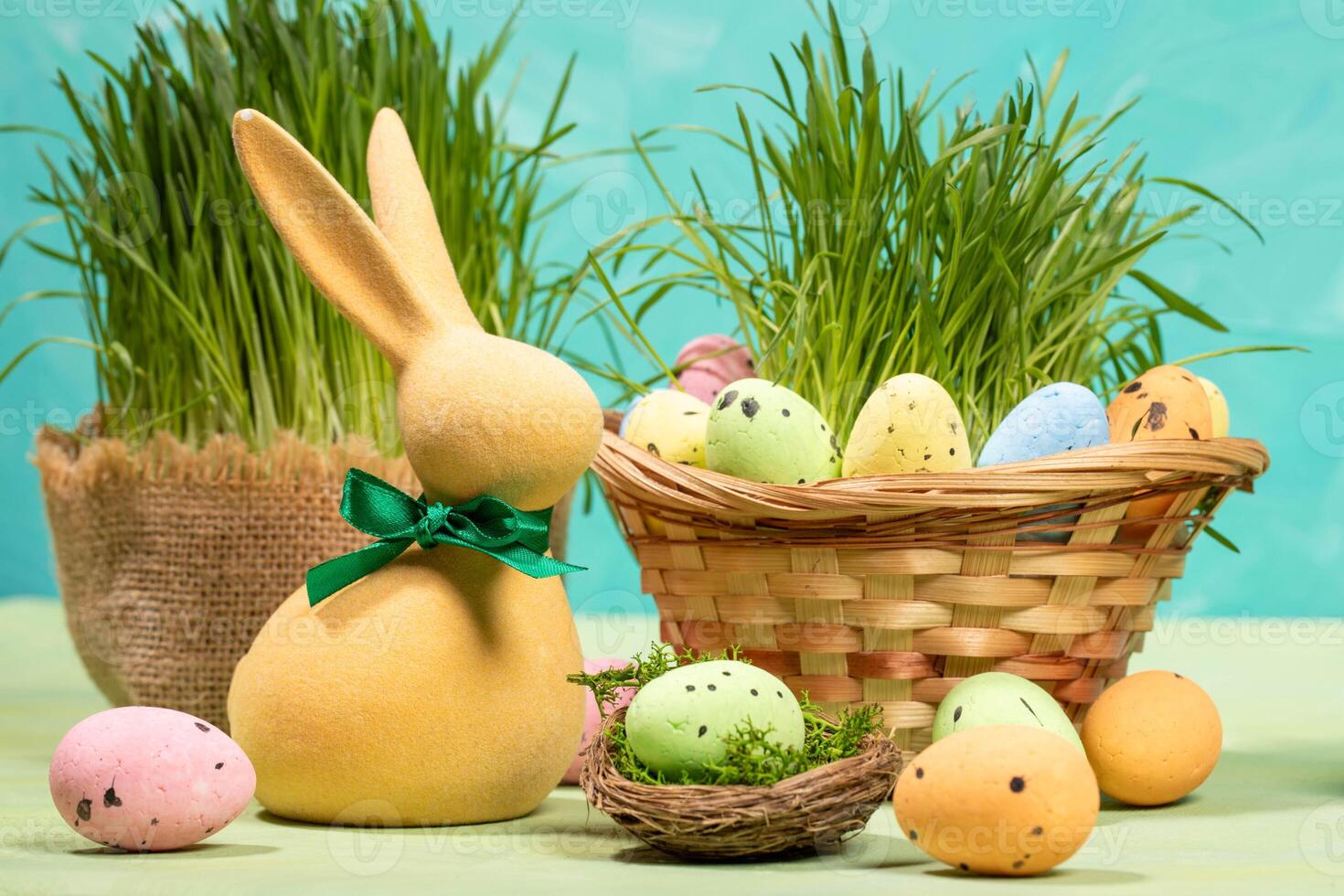 Easter card. Yellow bunny, colorful eggs on table, in nest, in basket with green grass on turquoise. photo
