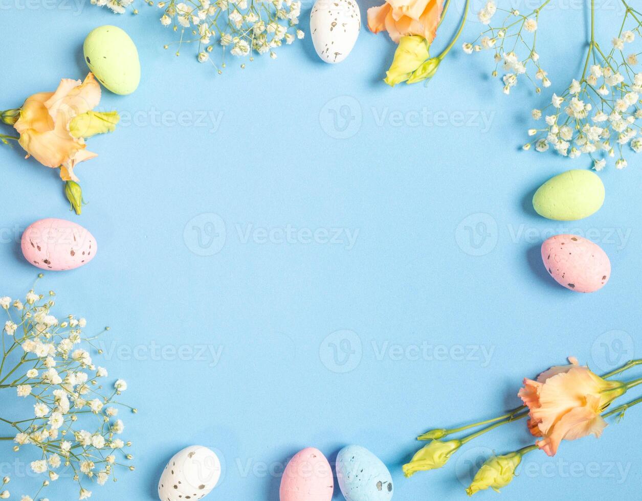 Frame of decorative Easter eggs and delicate white and peach flowers on blue. Top view. Copy space. photo