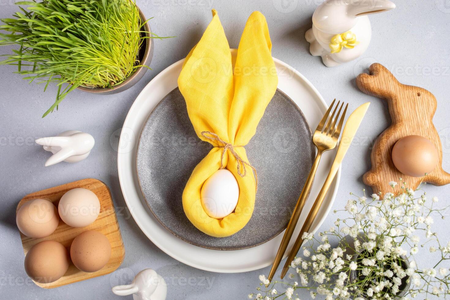 Napkin as bunny ears, plates, organic Easter eggs, grass, bunny, gold cutlery on gray. Top view. photo