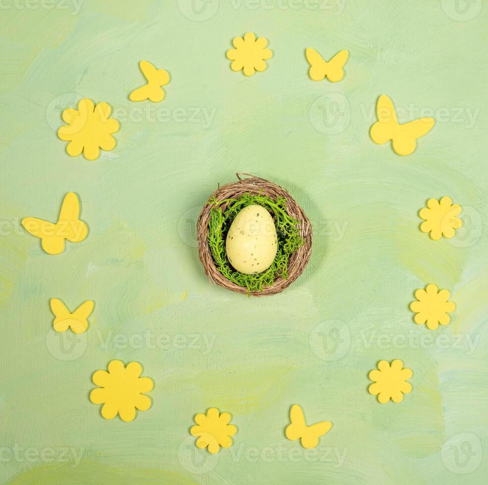 Easter egg in nest inside of round frame of yellow flowers and butterflies on green yellow. Top view photo