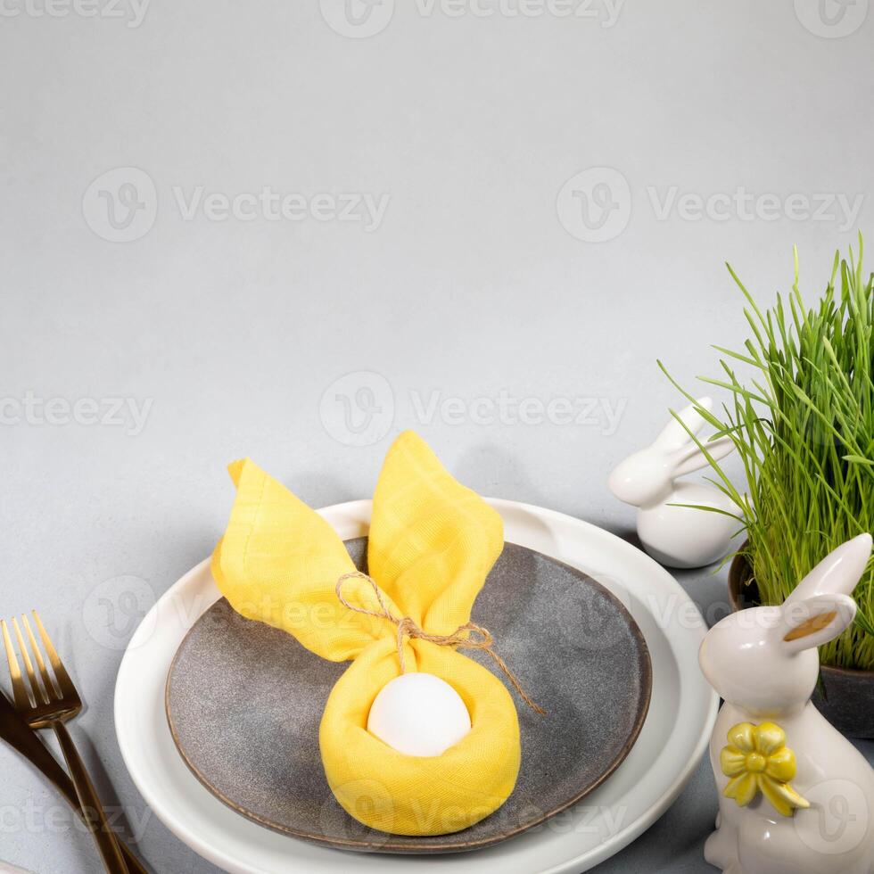 Plates, white Easter egg in yellow napkin, grass, ceramic bunnies, gold cutlery on gray. Copy space. photo