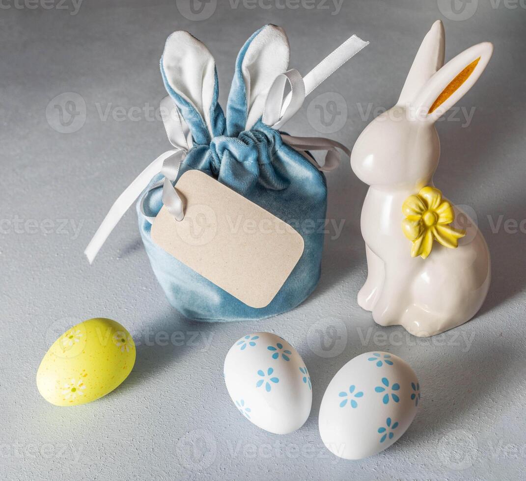 Easter image. Creative gift bag with bunny ears, blank note, ceramic bunny, decorative eggs on gray. photo