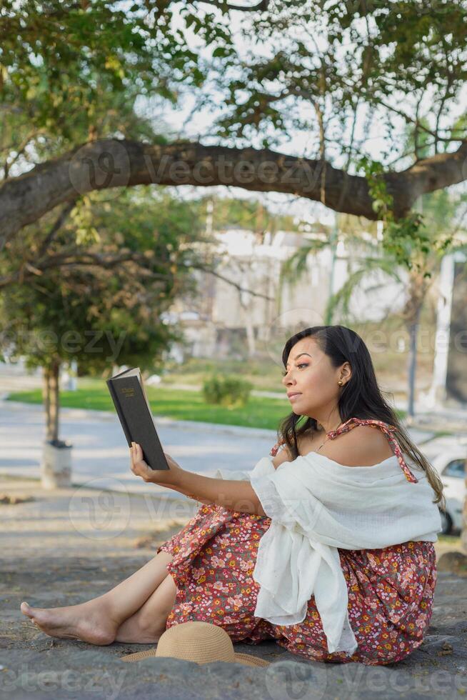 Woman wearing hat and dress with flowers, barefoot sitting on the stairs of a public park reading a book. photo