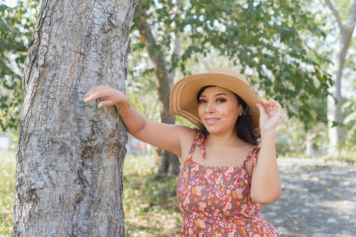 mujer vistiendo sombrero y vestir con flores admirativo naturaleza en un público parque cerca a un árbol. soleado verano día. foto