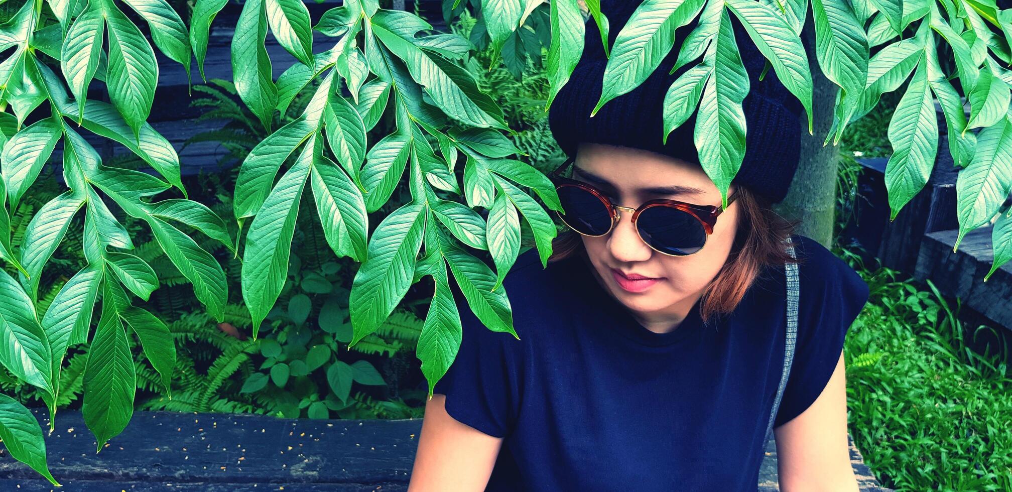 Asian woman wearing sunglass and wool hat sitting on wooden bench under tree with green plant background. Girl in casual dress style in vintage tone color with copy space. photo