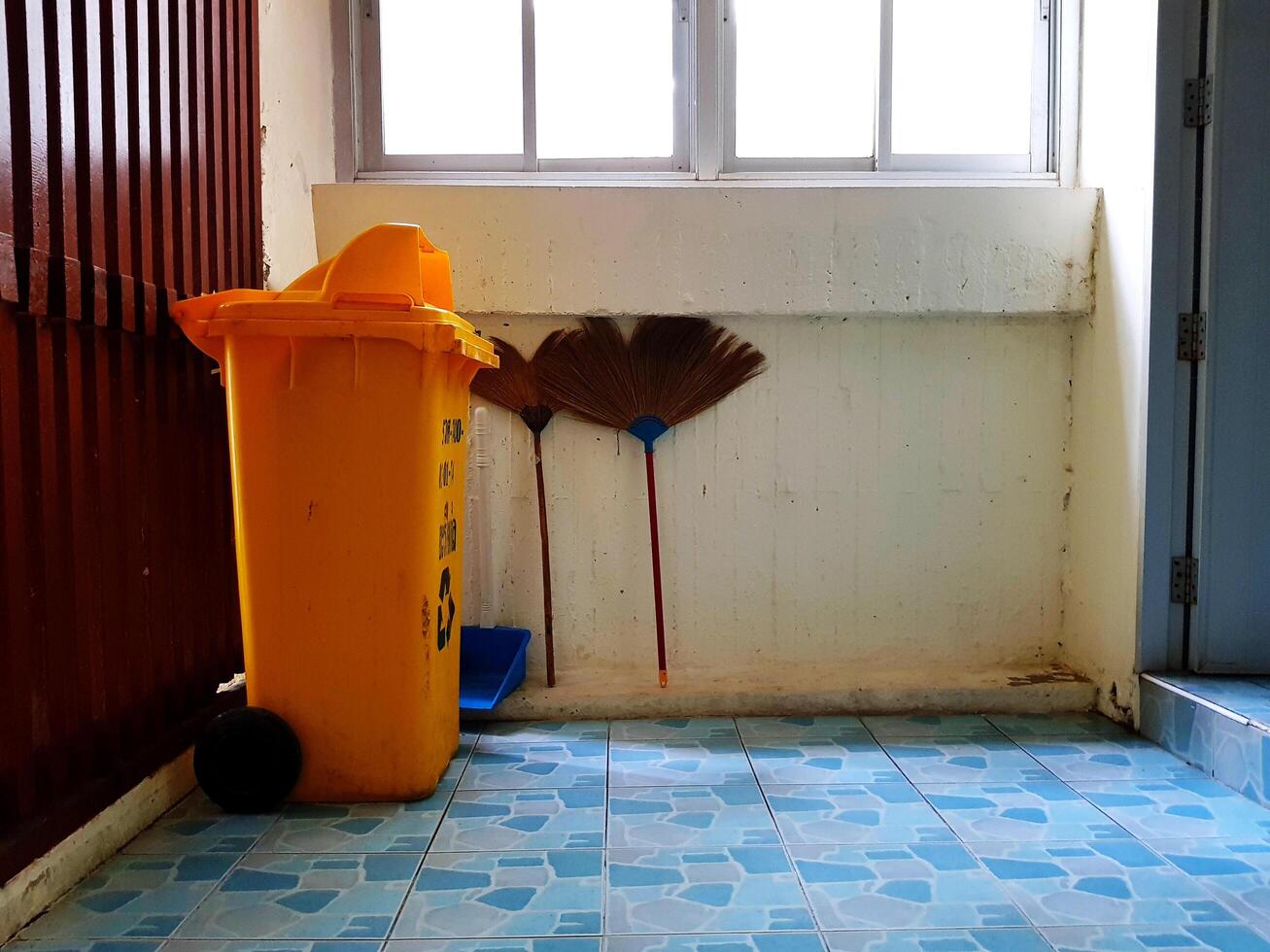 Yellow trash and broom placed on the blue tile floor with white and wooden wall with copy space. Cleaning tool concept photo