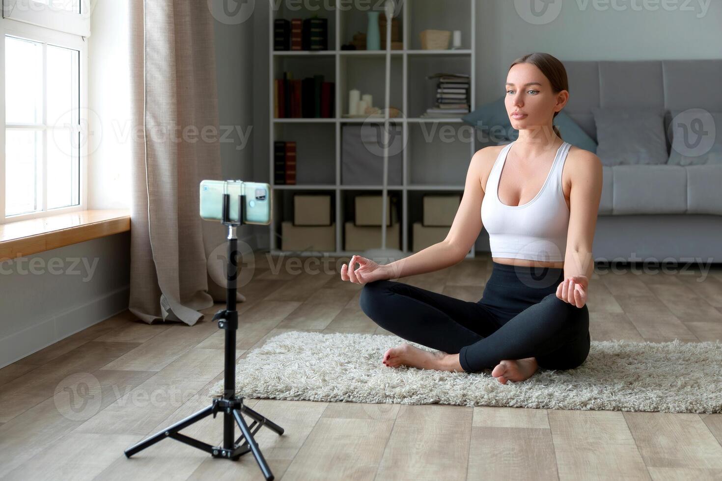 mujer haciendo yoga a hogar en el alfombra, sentado en loto posición con su teléfono y trípode para grabación s o En Vivo transmisión para un en línea aptitud centrar clase mediante un social medios de comunicación plataforma. foto