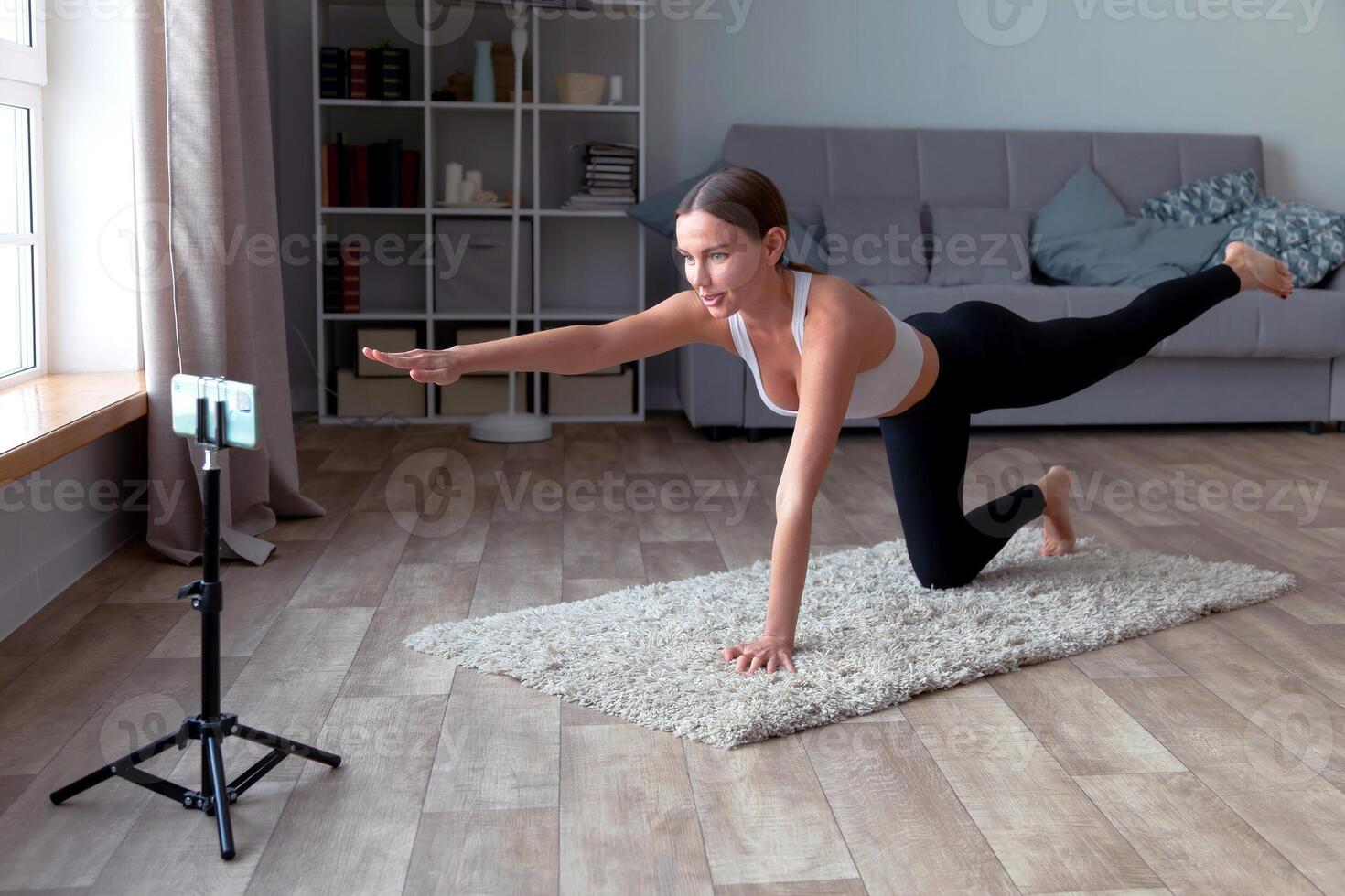 woman in black leggings and a white top does yoga at home on the carpet, recording an online fitness for her audience with a smartphone standing on a tripod photo
