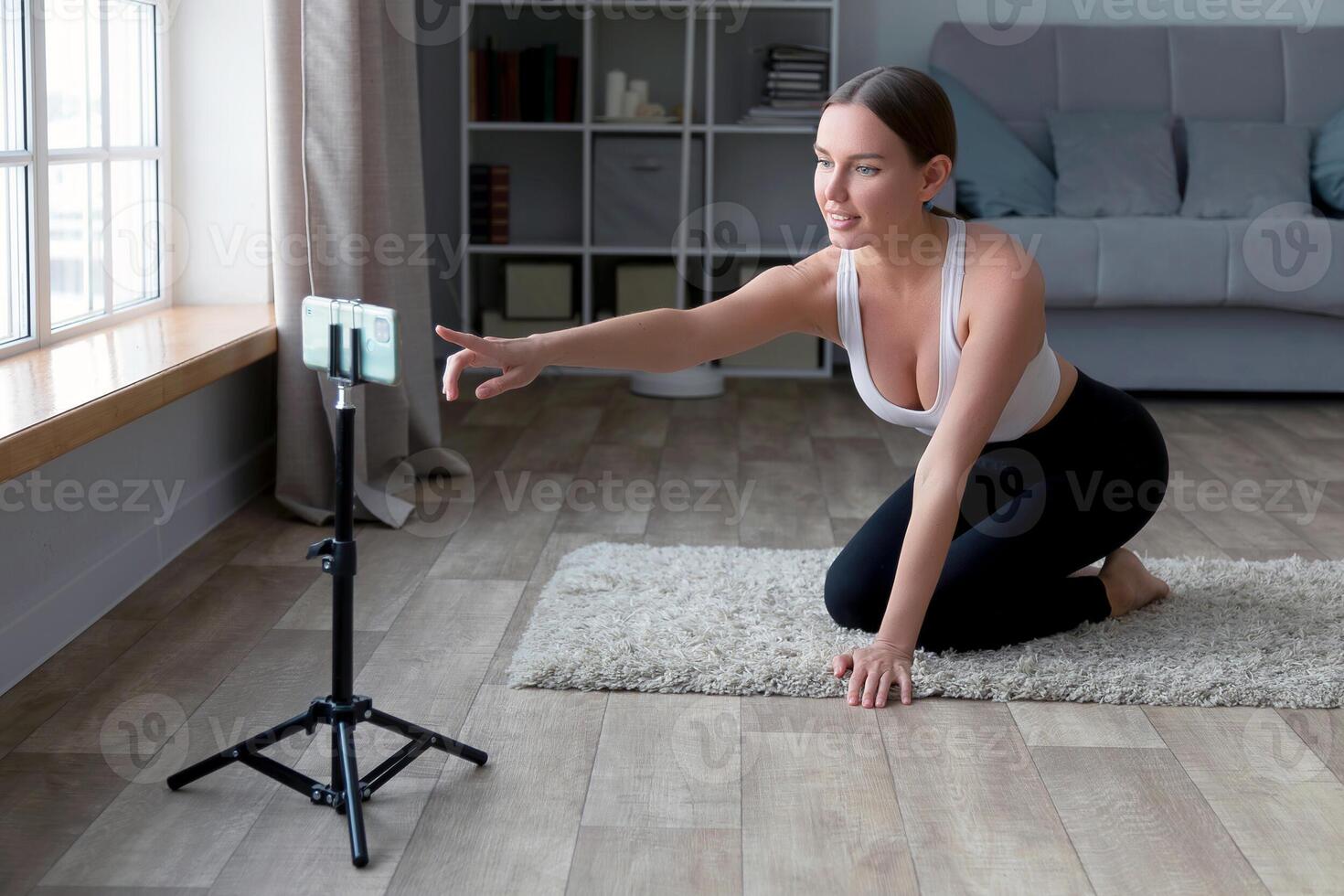 woman sitting on the floor in yoga pants recording an online fitness with her phone and tripod, smiling at the camera in a living room setting photo