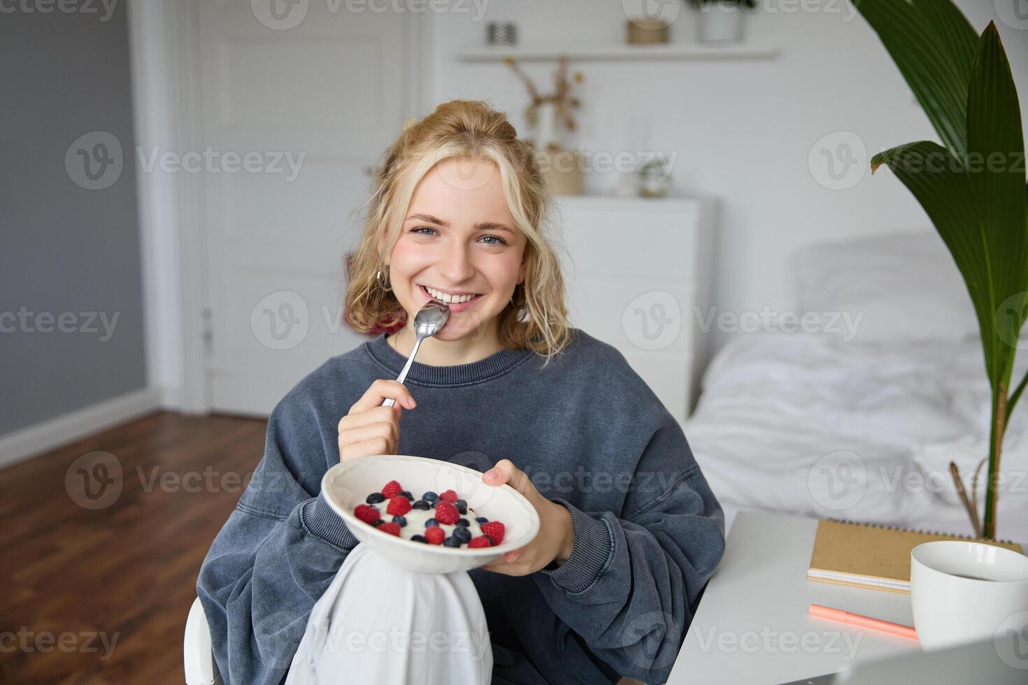Portrait of beautiful young woman in a room, eating breakfast, holding bowl with dessert and a spoon, smiling at camera, recording lifestyle vlog photo