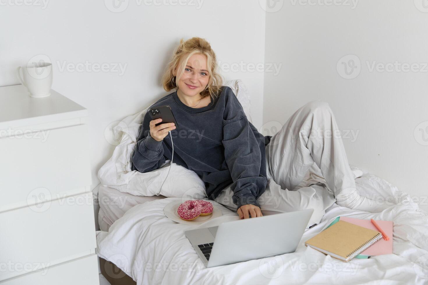 Portrait of young woman, student studying in her bed, relaxing while preparing homework, eating doughnut, using laptop in bedroom and drinking tea photo