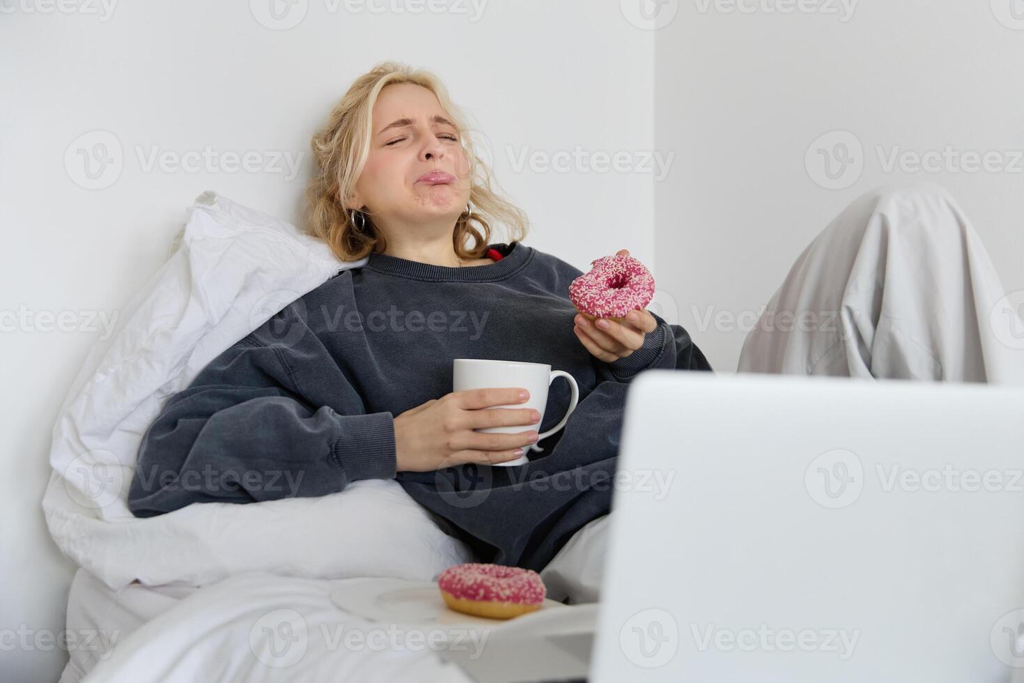 Portrait of sad and depressed woman, feeling heartbroken, lying in bed with comfort food, eating doughnut, crying from upsetting movie, watching something on laptop photo