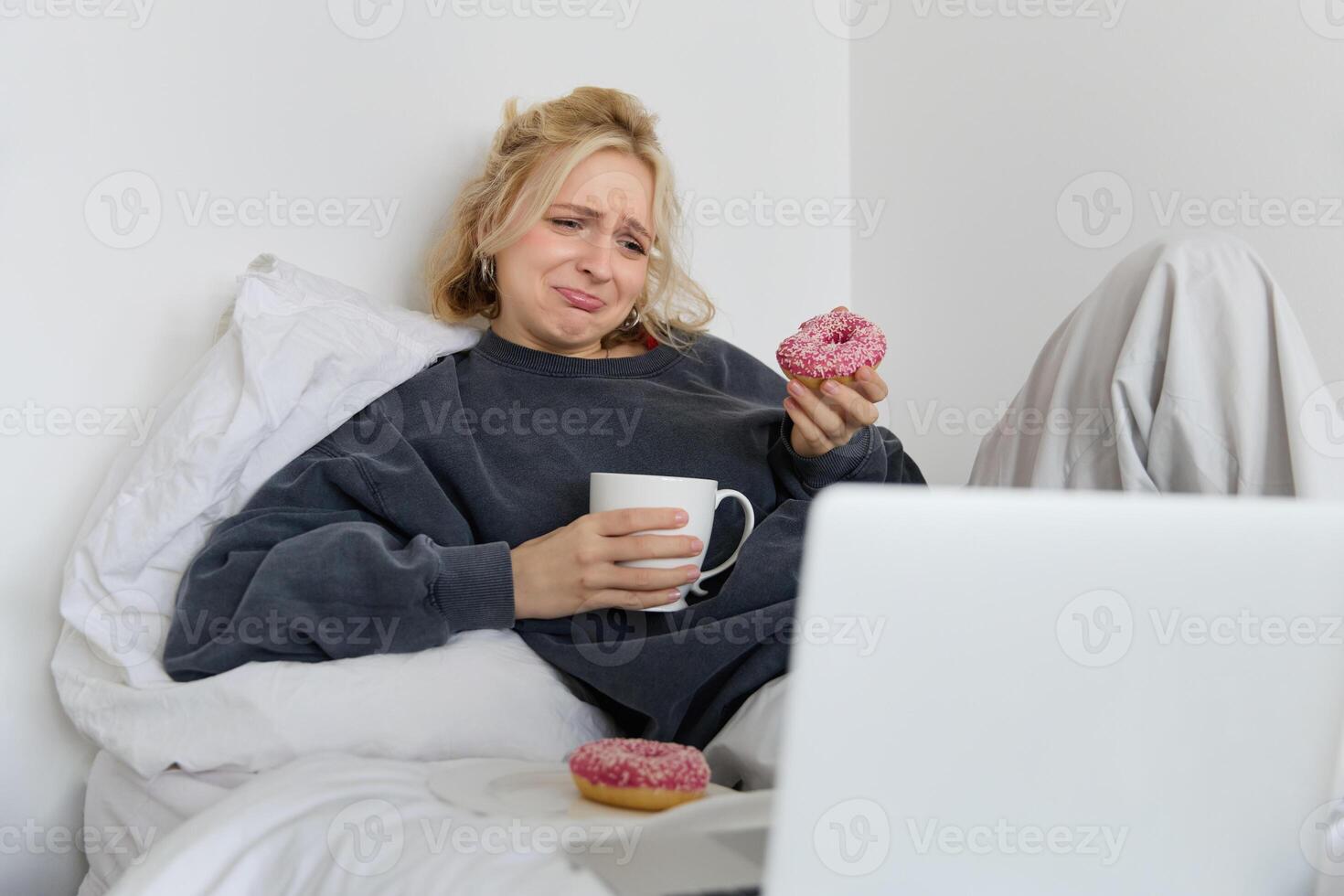 Portrait of sad, crying young woman, staying at home, sitting in bed with doughnut and comfort food, looking at something upsetting on laptop screen photo