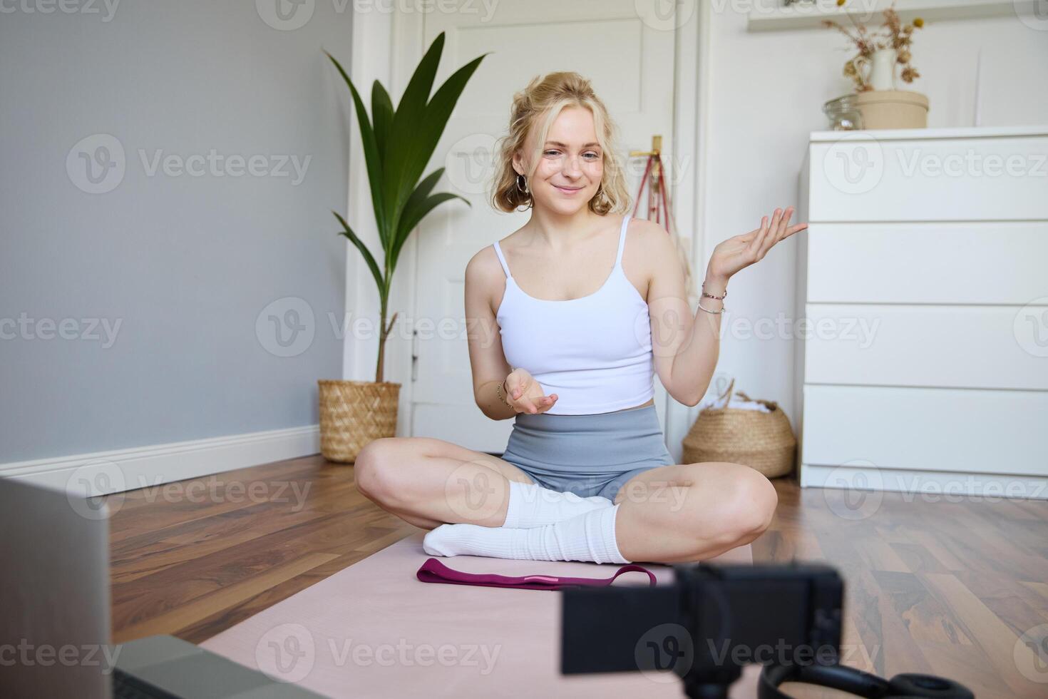 Portrait of beautiful fitness blogger, recording on digital camera, showing workout exercises, explaining fitness movements or yoga training to followers, sits on rubber mat photo
