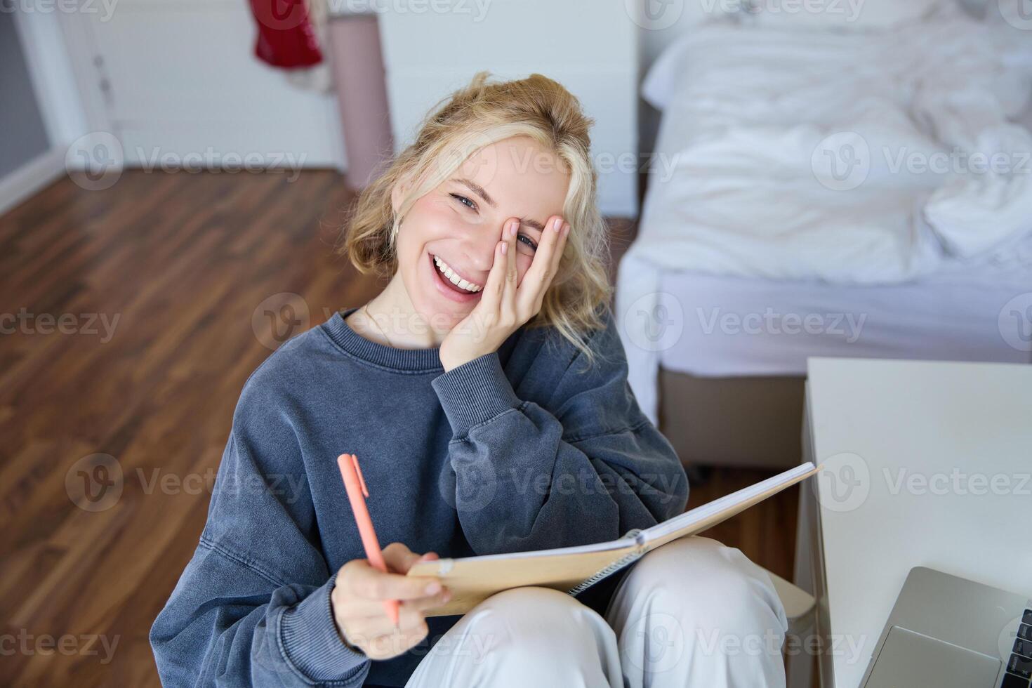 Portrait of happy, young blond woman, sitting in bedroom with notebook and pen, laughing and smiling, writing in journal, doing homework and smiling photo