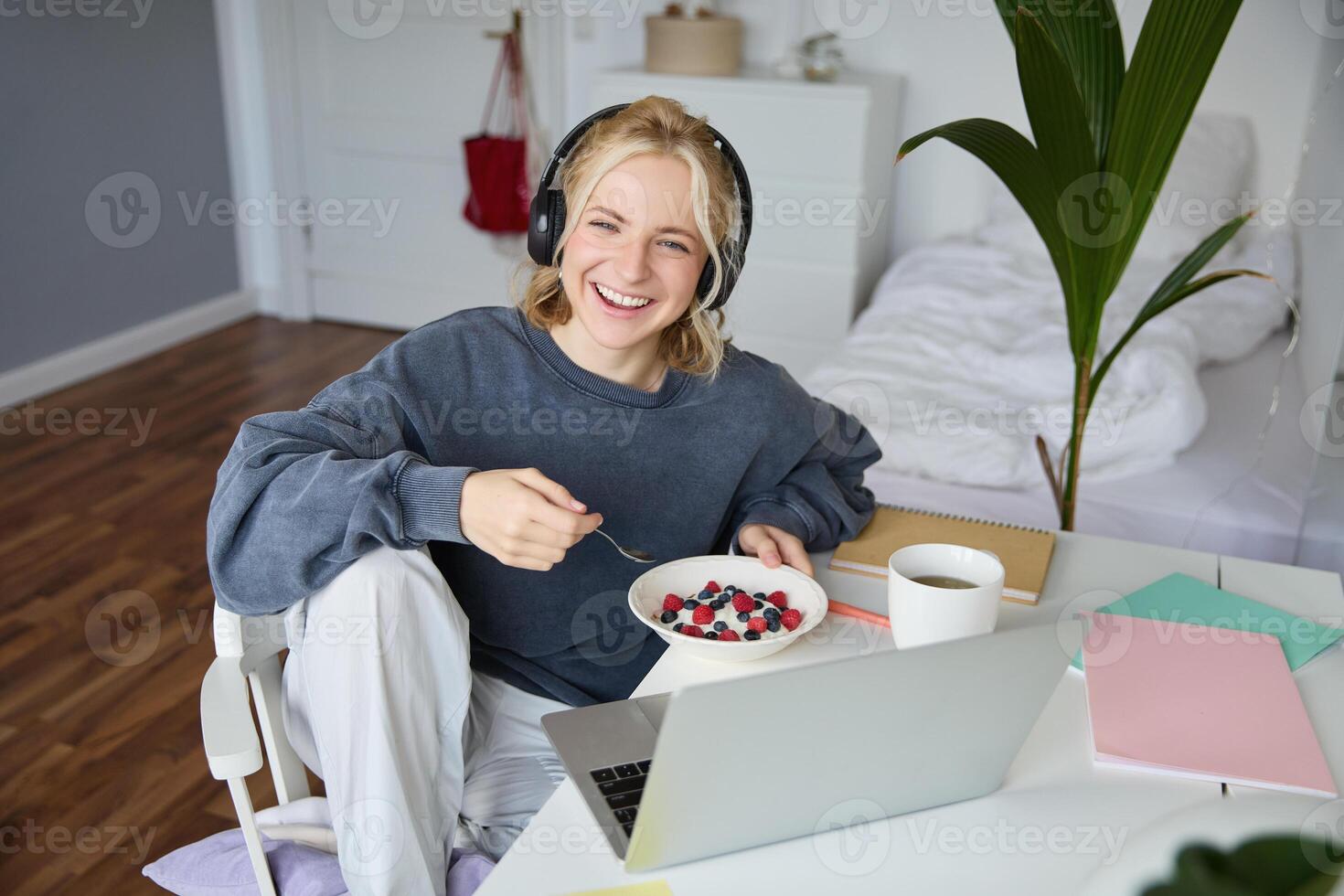 Image of happy woman sitting in a room, watching interesting tv show or movie on laptop, using screaming service, wearing headphones, eating dessert and drinking tea photo